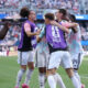 Soccer players in purple bibs celebrate together on the field as fans watch from the stands behind them.