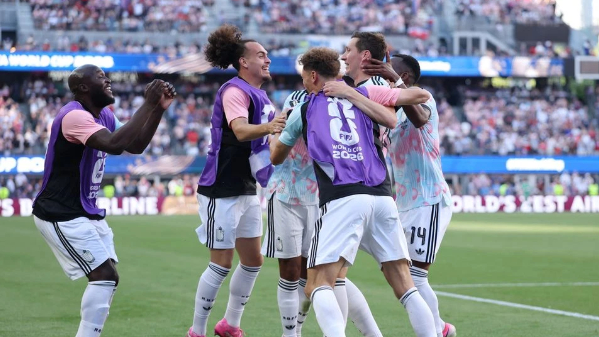 Soccer players in purple bibs celebrate together on the field as fans watch from the stands behind them.