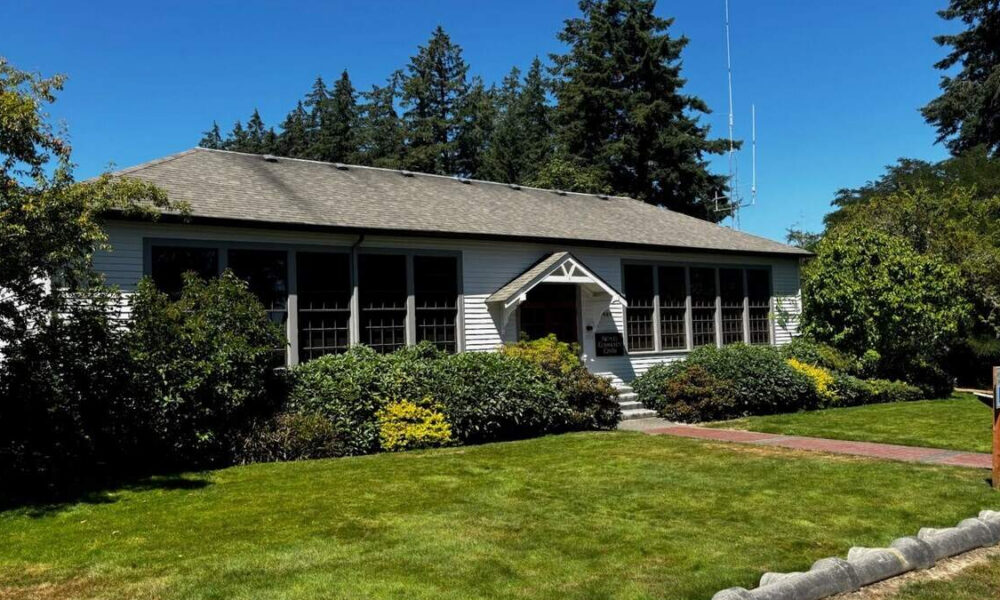 Small white, single-story building with a gabled entrance, shrubs, and a green lawn on a sunny day.