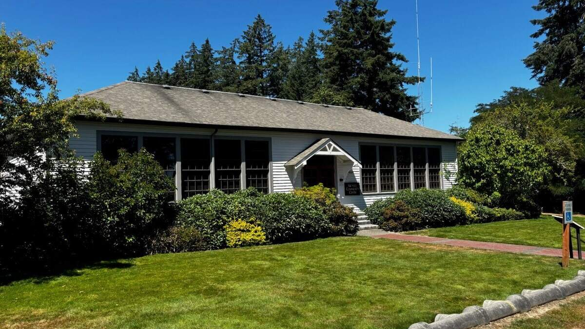 Small white, single-story building with a gabled entrance, shrubs, and a green lawn on a sunny day.