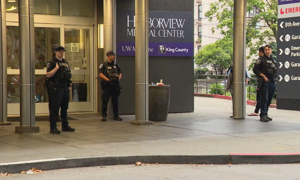 Four police officers in tactical gear stand guard outside Harborview Medical Center entrance and signage visible above.