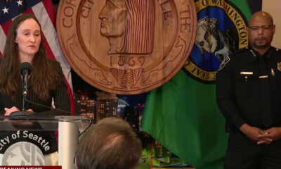 Woman speaks at a City of Seattle press conference podium, with American flag and large seal backdrop; a police officer stands to the right.