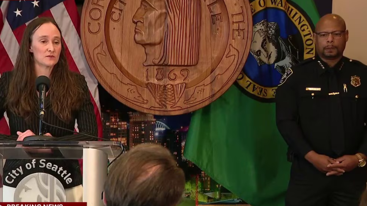 Woman speaks at a City of Seattle press conference podium, with American flag and large seal backdrop; a police officer stands to the right.