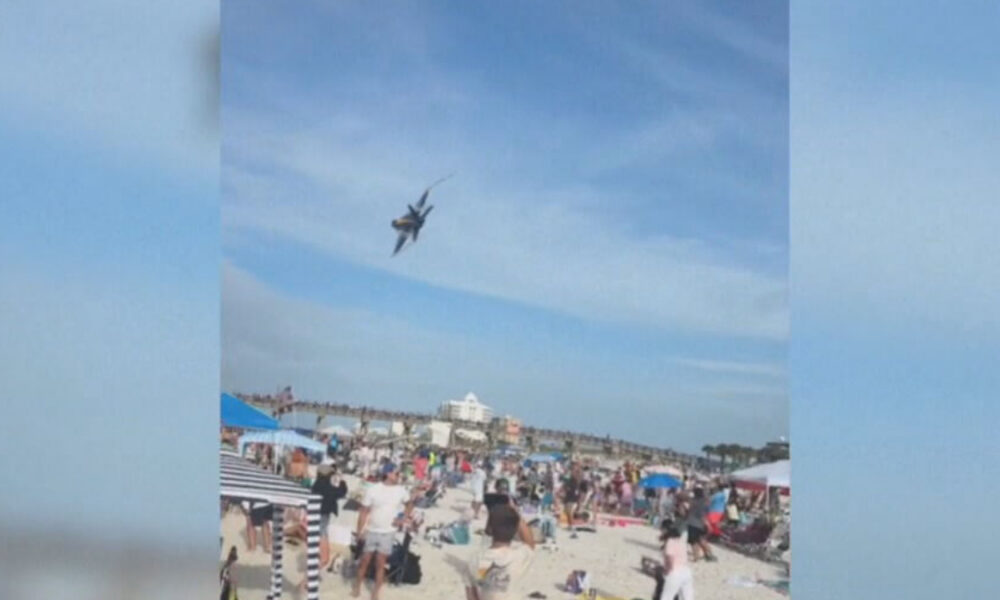 Jet performing a low pass over a crowded beach festival with people sunbathing and umbrellas visible at the shore below.