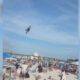 Jet performing a low pass over a crowded beach festival with people sunbathing and umbrellas visible at the shore below.