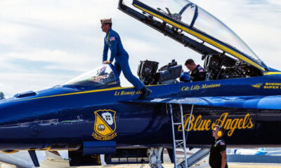 Blue Angels fighter jet with open canopy; a crew member in a blue flight suit climbs into the cockpit while ground crew members observe nearby.