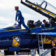 Blue Angels fighter jet with open canopy; a crew member in a blue flight suit climbs into the cockpit while ground crew members observe nearby.