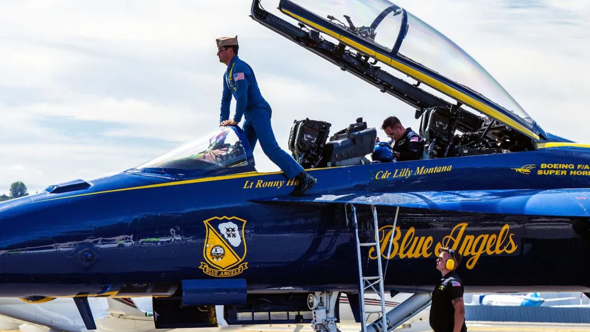 Blue Angels fighter jet with open canopy; a crew member in a blue flight suit climbs into the cockpit while ground crew members observe nearby.