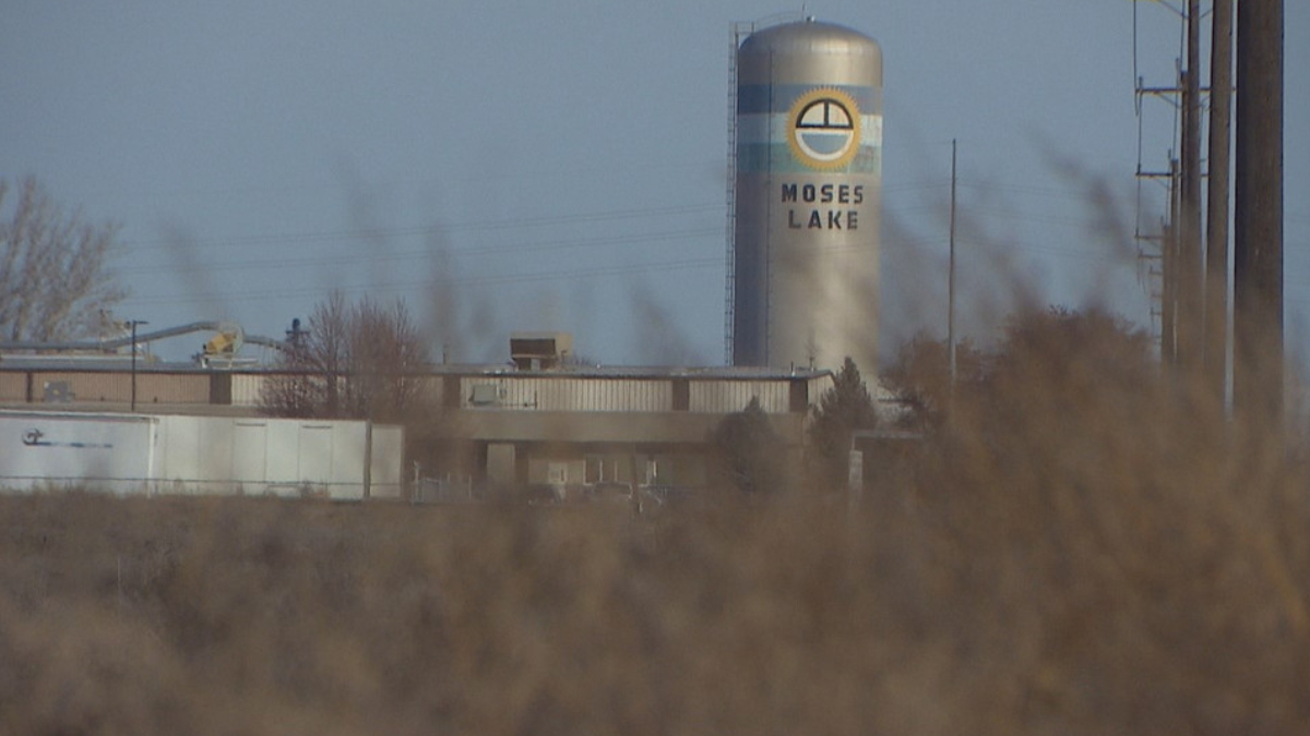 Tall silo with Moses Lake branding behind low industrial buildings, with dry brush in the foreground.