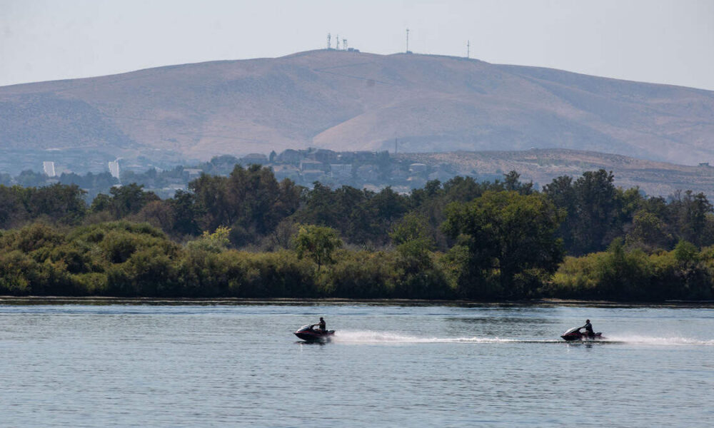 Two people ride jet skis across a calm river, with a forested shoreline and a hill with antennas in the distance.