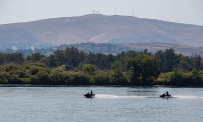 Two people ride jet skis across a calm river, with a forested shoreline and a hill with antennas in the distance.