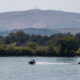 Two people ride jet skis across a calm river, with a forested shoreline and a hill with antennas in the distance.