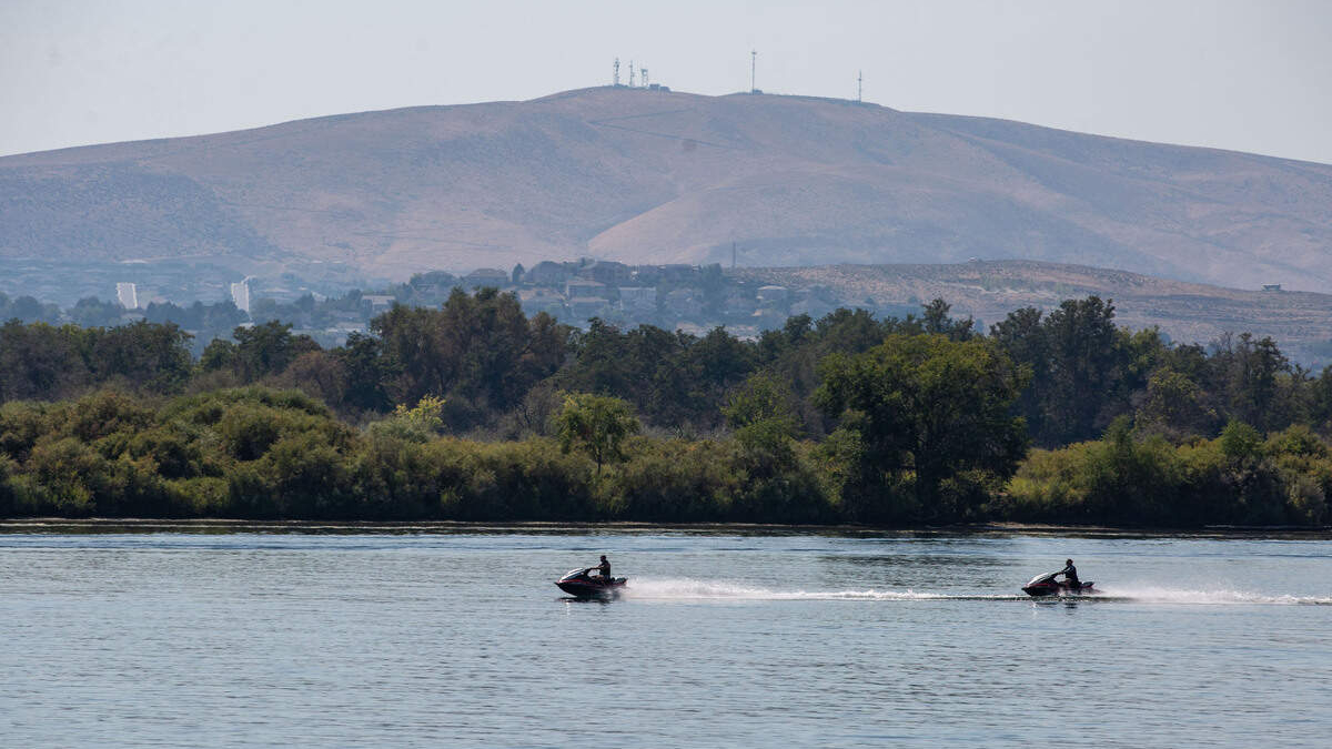 Two people ride jet skis across a calm river, with a forested shoreline and a hill with antennas in the distance.