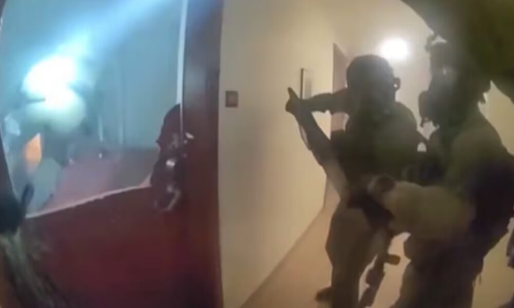 A group of soldiers in tactical gear with rifles advancing down a hallway toward a doorway, ready for entry.