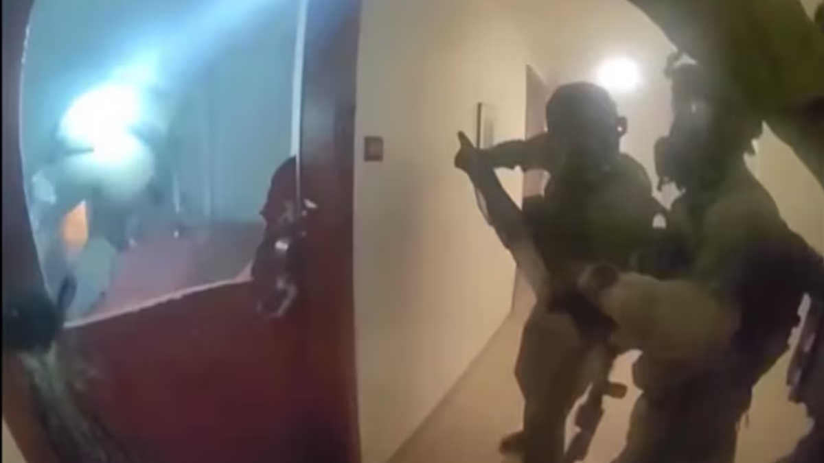 A group of soldiers in tactical gear with rifles advancing down a hallway toward a doorway, ready for entry.