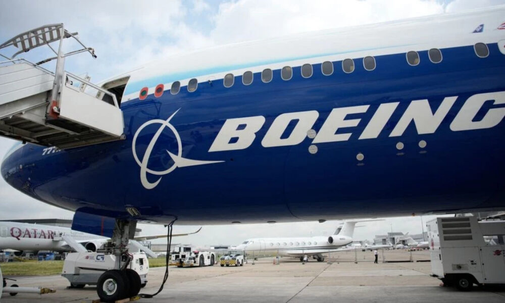 Blue Boeing passenger jet at the gate with a jet bridge connected and other aircraft on the tarmac in the background.