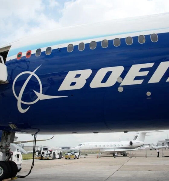 Blue Boeing passenger jet at the gate with a jet bridge connected and other aircraft on the tarmac in the background.