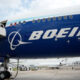 Blue Boeing passenger jet at the gate with a jet bridge connected and other aircraft on the tarmac in the background.