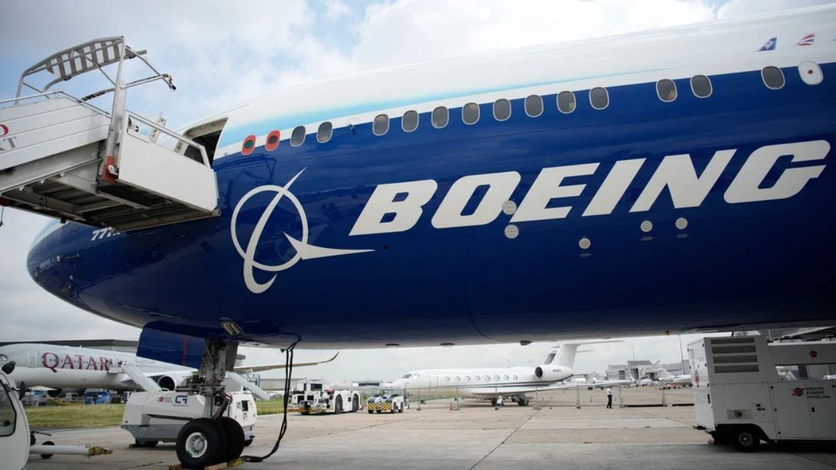 Blue Boeing passenger jet at the gate with a jet bridge connected and other aircraft on the tarmac in the background.