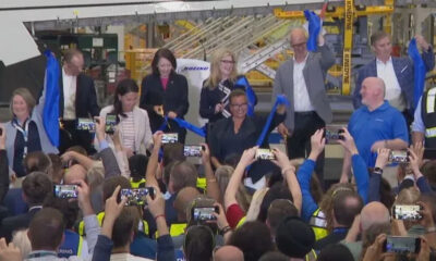 Executives and staff cutting a blue ribbon at a launch event inside an aircraft manufacturing facility after a ceremony, crowd filming with smartphones nearby