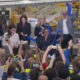 Executives and staff cutting a blue ribbon at a launch event inside an aircraft manufacturing facility after a ceremony, crowd filming with smartphones nearby