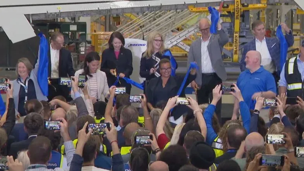 Executives and staff cutting a blue ribbon at a launch event inside an aircraft manufacturing facility after a ceremony, crowd filming with smartphones nearby