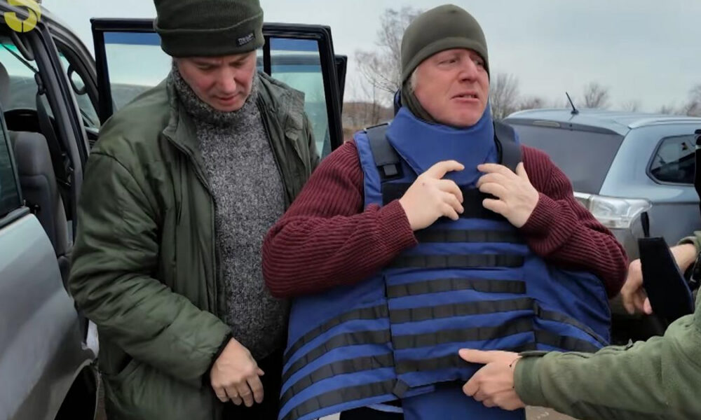 Two men in a cold outdoor parking area; one in a green jacket helps another man wearing a blue protective vest and a gray beanie, with a Ukrainian flag in the background.