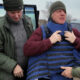 Two men in a cold outdoor parking area; one in a green jacket helps another man wearing a blue protective vest and a gray beanie, with a Ukrainian flag in the background.
