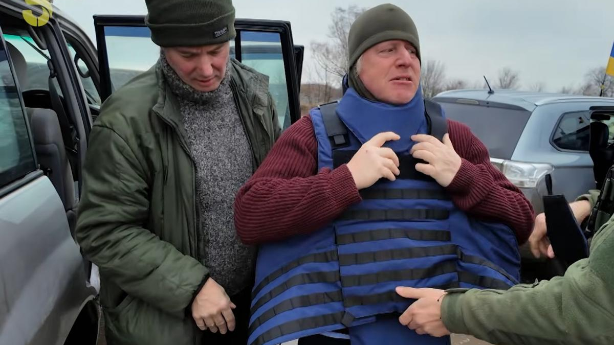 Two men in a cold outdoor parking area; one in a green jacket helps another man wearing a blue protective vest and a gray beanie, with a Ukrainian flag in the background.