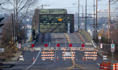 Wide shot of a street blocked by orange-and-white barricades and orange cones, sign reading ROAD CLOSED across the road, with a green metal bridge behind.