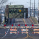 Wide shot of a street blocked by orange-and-white barricades and orange cones, sign reading ROAD CLOSED across the road, with a green metal bridge behind.