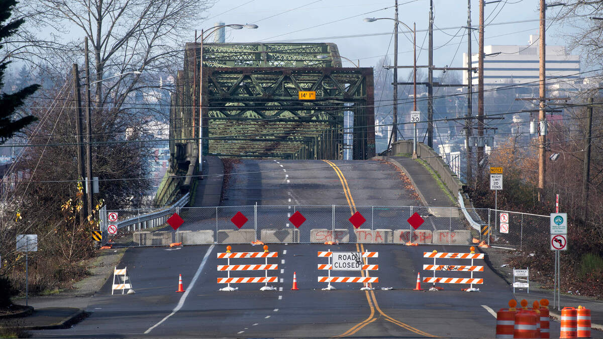 Wide shot of a street blocked by orange-and-white barricades and orange cones, sign reading ROAD CLOSED across the road, with a green metal bridge behind.