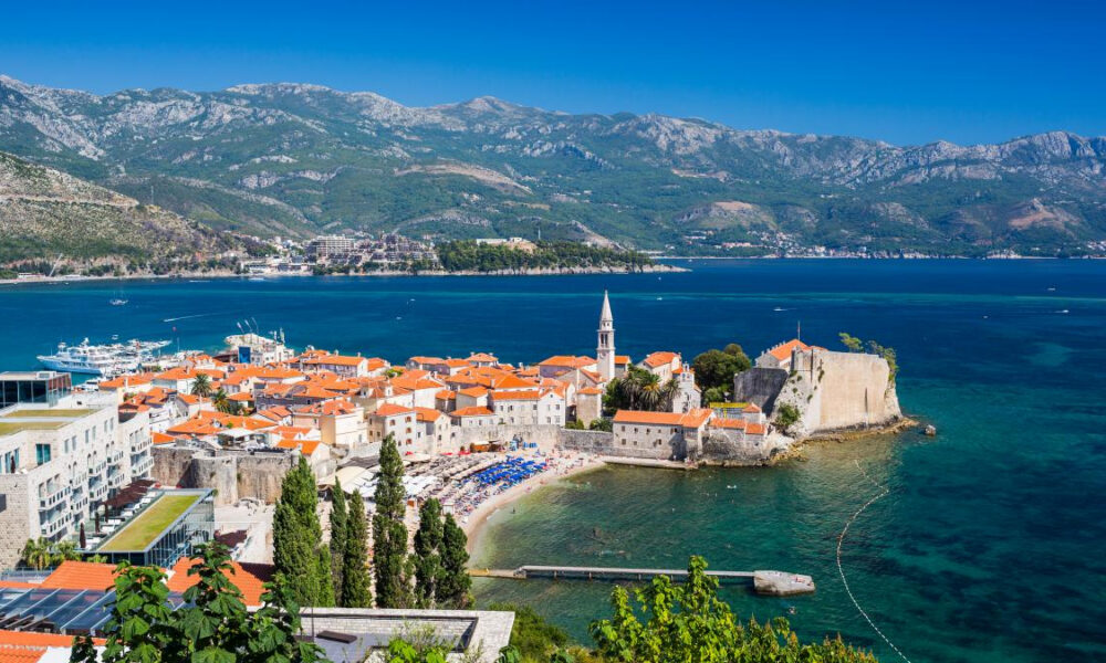 Coastal town with red-roofed houses, a medieval fortress, and a church steeple along a blue bay and distant mountains.