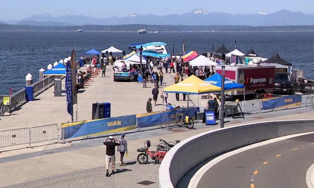 Crowd at a seaside festival on a pier with colorful tents, banners, and boats on the water.
