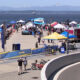 Crowd at a seaside festival on a pier with colorful tents, banners, and boats on the water.