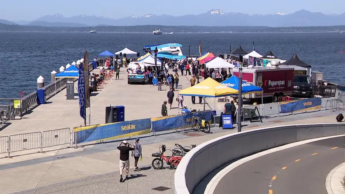 Crowd at a seaside festival on a pier with colorful tents, banners, and boats on the water.