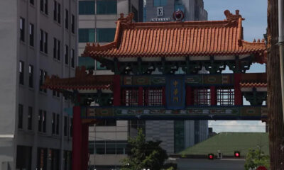 Ornate Chinese-style gate with red columns and orange tiled roof spanning a city street between office buildings, blue sign with gold Chinese characters.