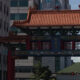 Ornate Chinese-style gate with red columns and orange tiled roof spanning a city street between office buildings, blue sign with gold Chinese characters.