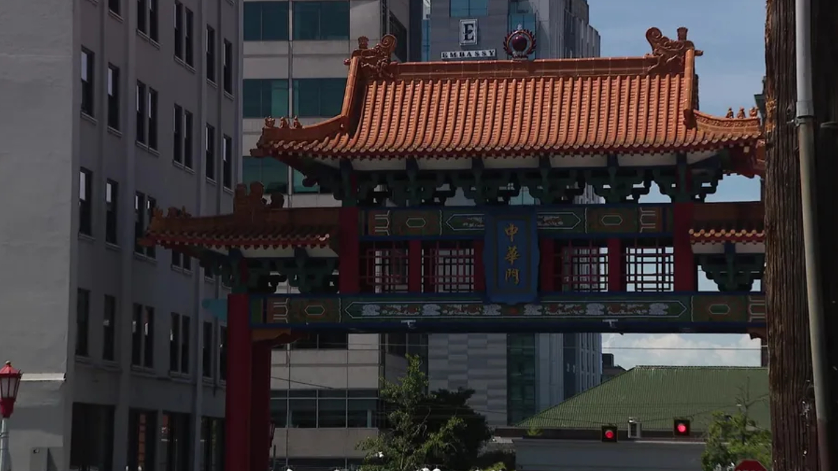 Ornate Chinese-style gate with red columns and orange tiled roof spanning a city street between office buildings, blue sign with gold Chinese characters.