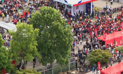 Bird's-eye view of a crowded outdoor festival with red tents, banners, and groups of people among green trees, seen from above.