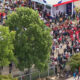 Bird's-eye view of a crowded outdoor festival with red tents, banners, and groups of people among green trees, seen from above.