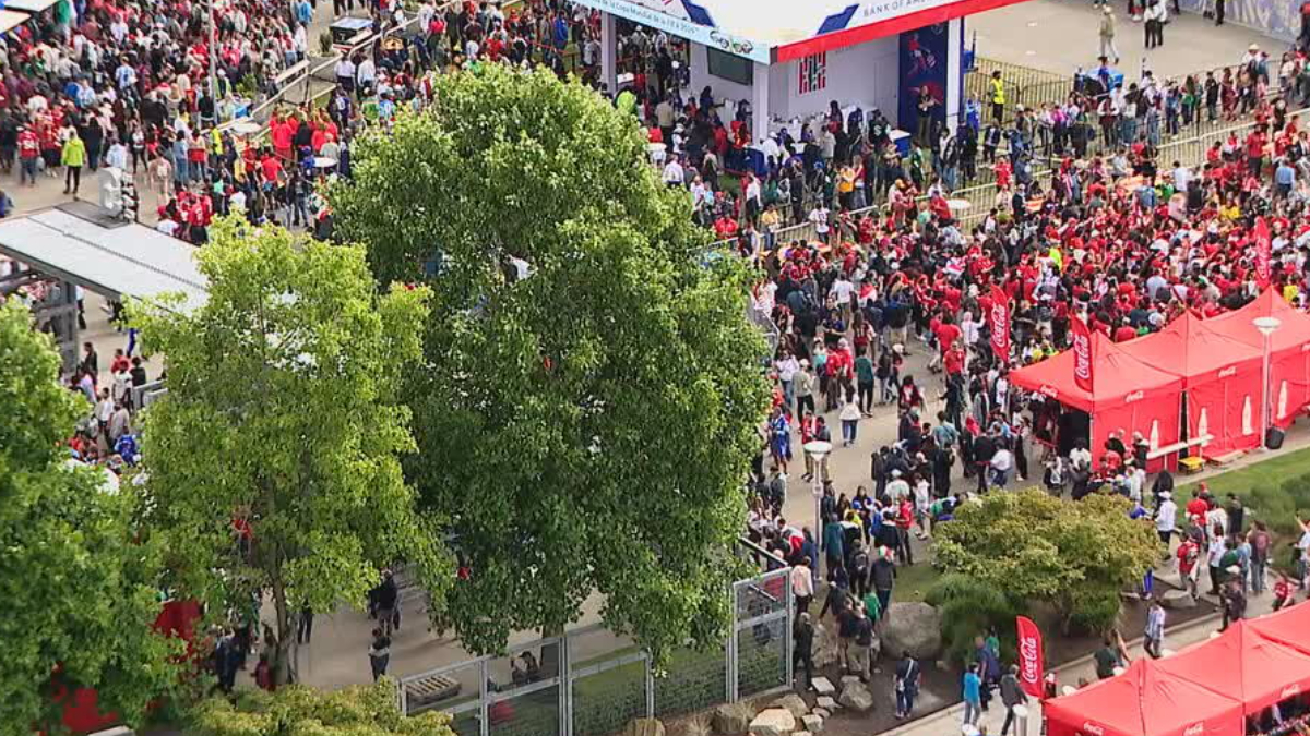 Bird's-eye view of a crowded outdoor festival with red tents, banners, and groups of people among green trees, seen from above.