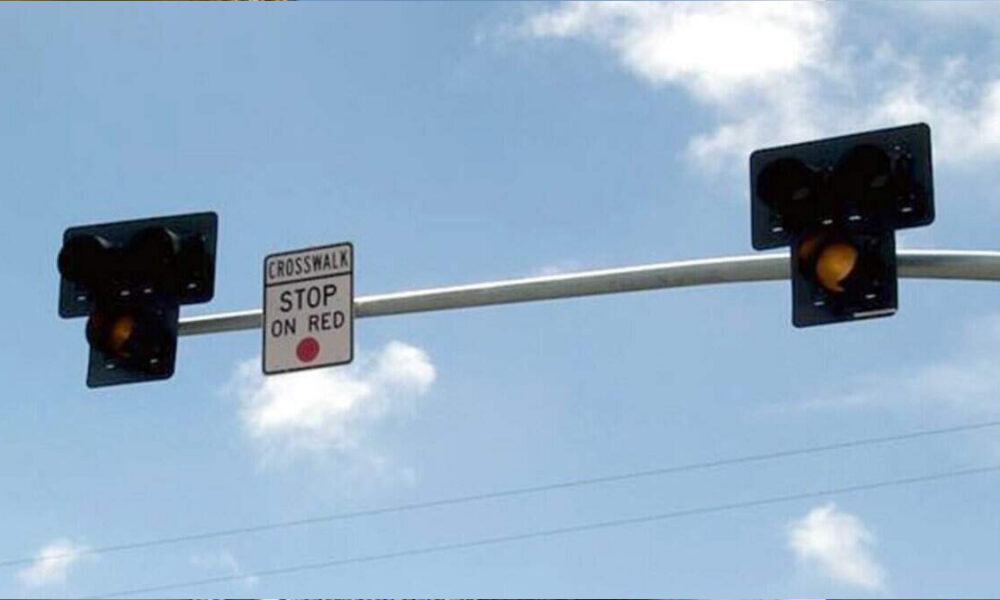 Two traffic lights mounted on a pole with a small 'Crosswalk Stop on Red' sign between them against a blue sky backdrop, warning drivers to stop on red.