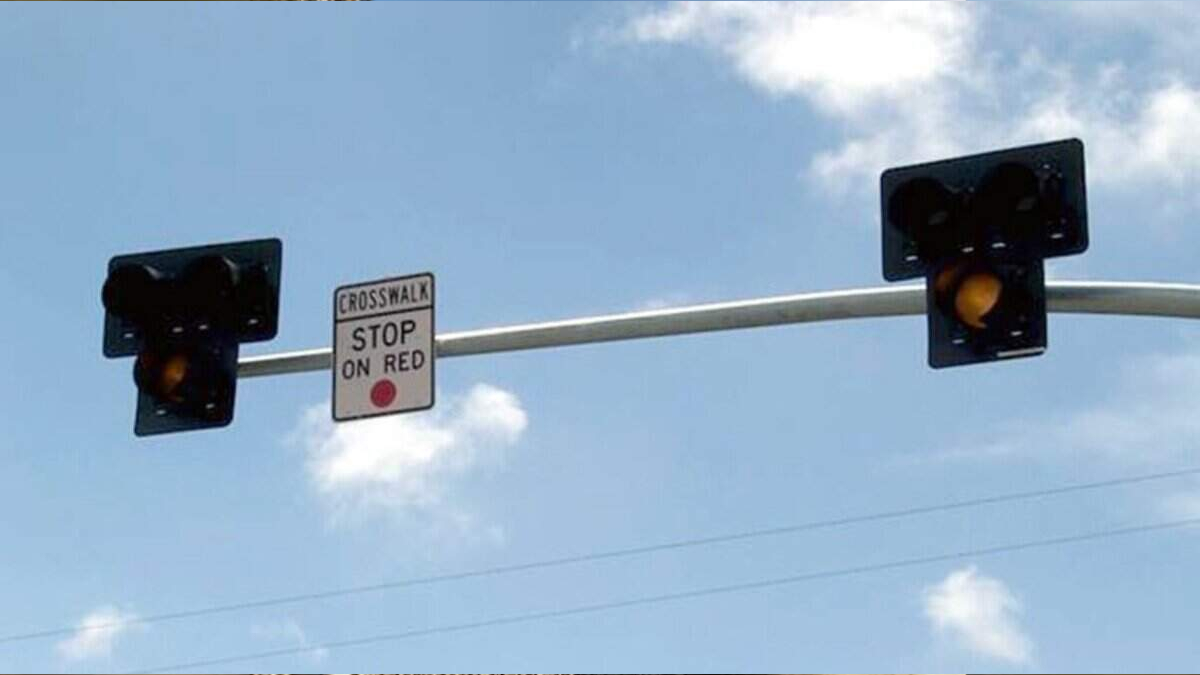 Two traffic lights mounted on a pole with a small 'Crosswalk Stop on Red' sign between them against a blue sky backdrop, warning drivers to stop on red.