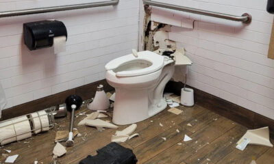 Damaged bathroom with a toilet and a large hole in the wall behind it, debris scattered on the floor.