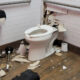 Damaged bathroom with a toilet and a large hole in the wall behind it, debris scattered on the floor.