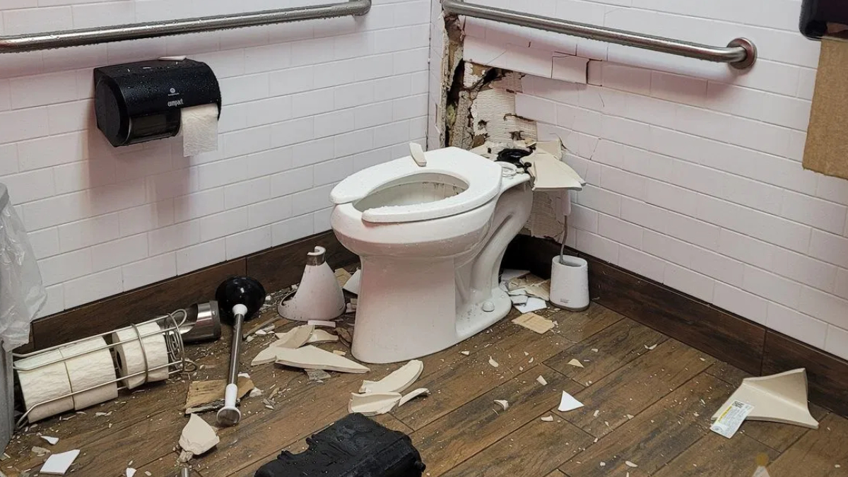 Damaged bathroom with a toilet and a large hole in the wall behind it, debris scattered on the floor.