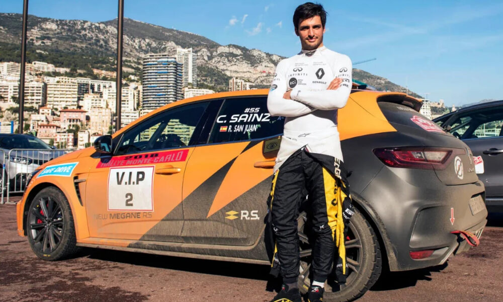 Racing driver in white suit stands with folded arms beside an orange VIP‑2 race car, city skyline behind them.