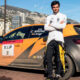 Racing driver in white suit stands with folded arms beside an orange VIP‑2 race car, city skyline behind them.