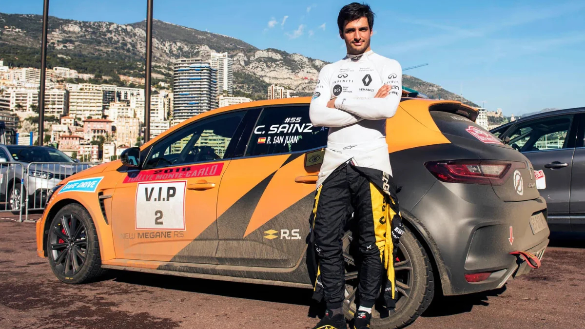 Racing driver in white suit stands with folded arms beside an orange VIP‑2 race car, city skyline behind them.